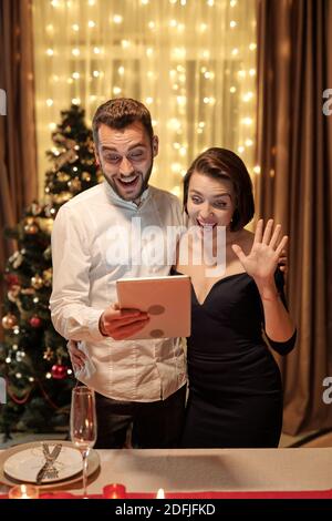 Giovane coppia allegra ed elegante che guarda lo schermo dello smartphone mentre comunica a qualcuno in video chat e moglie che agita mano Foto Stock