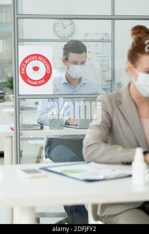 Giovane uomo d'affari in maschera protettiva e abbigliamento da formalwear guardando il computer portatile visualizzazione quando si lavora con i dati in ufficio davanti a. collega Foto Stock