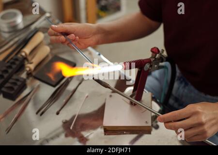 Mani di giovane lavoratrice femminile che tiene due pezzi sopra il bruciatore durante la preparazione per l'ulteriore lavorazione in officina o in studio Foto Stock