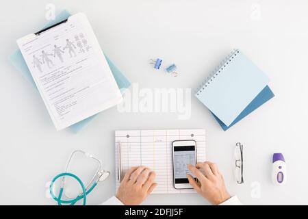 Panoramica delle mani del medico sulla scheda vuota e sul touchscreen di gadget mentre si scorrono i dati nello smartphone in base al luogo di lavoro Foto Stock