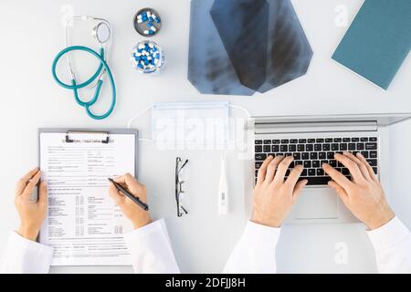 Flatlay di mani di medici di sesso maschile e femminile che fanno note mediche e la digitazione da computer portatile circondato da immagini radiografiche, pillole e stetoscopio Foto Stock