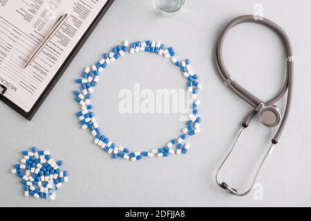 Pillole blu e bianche che formano un grande cerchio sul posto di lavoro con vetro di acqua, stetoscopio, documento con penna e pila di capsule intorno Foto Stock