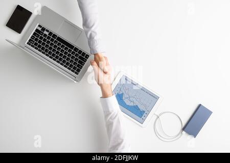 Vista dall'alto delle mani di due giovani contemporanei che salutano ciascuno altro da handshake tra i gadget mobili su sfondo bianco Foto Stock
