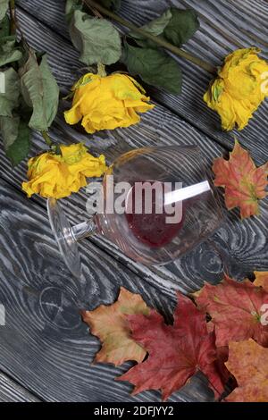 Un bouquet di rose gialle secche e foglie d'autunno secche. Un bicchiere rovesciato di vino affolto. Su tavole di pino. Foto Stock