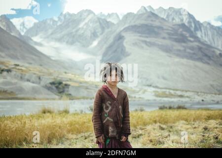 Ragazza, di 8 anni, in piedi davanti ad un campo di grano raccolto, membri del gruppo etnico del Wakhi stabilito, dietro di loro i piedi del Foto Stock