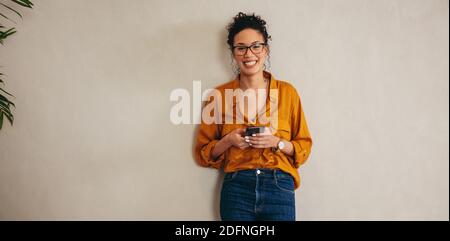 Ritratto di una giovane donna felice in piedi contro un muro. Donna imprenditore che tiene un telefono cellulare guardando la macchina fotografica e sorridendo. Foto Stock