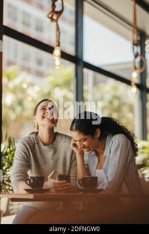 Due donne che guardano il telefono e ridono all'interno di una caffetteria. Gli amici seduti con le tazze di caffè sul tavolo si divertono. Foto Stock