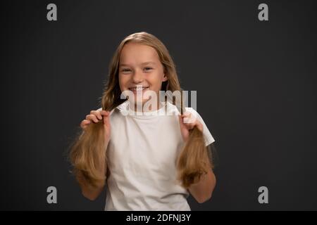 Sorridente tenendo due code di pony ragazzina. Indossare una t-shirt bianca sorridendo un po' di grilling sulla fotocamera isolata su sfondo grigio scuro o nero Foto Stock