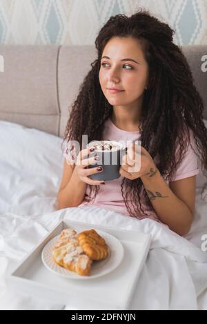 Le donne hanno la prima colazione a letto in un appartamento d'albergo leggero o a casa. Finestra ritratto luce giovane ragazza mangiando croissant e bevendo caffè. Foto Stock