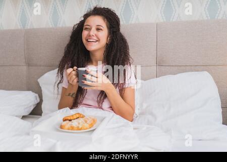 Le donne hanno la prima colazione a letto in un appartamento d'albergo leggero o a casa. Finestra luce ritratto giovane ragazza mangiando croissant, bevendo caffè e sorridendo. Foto Stock