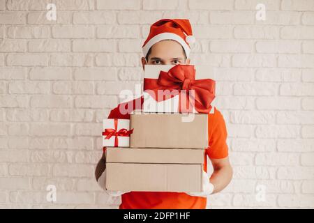 Divertente uomo di consegna in cappello di Santa, maschera medica viso, guanti di protezione portare scatole regalo di cartone con arco rosso. Felice corriere giovane consegna Christm Foto Stock