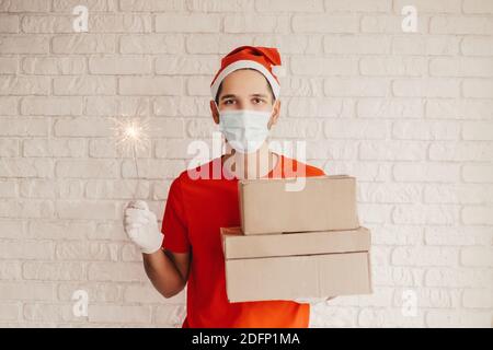 Giovane addetto al servizio di consegna in maschera protettiva, i guanti medici tengono scatole di cartone, scintilla in mani. Consegna felice ragazzo corriere in maschera medica Foto Stock