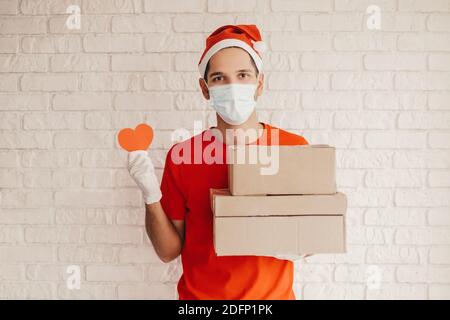 Felice ragazzo di consegna in maschera medica, guanti protettivi tenere confezioni di cartone, segno cuore in mano. Dipendente del servizio di consegna con scatole di spedizione. Foto Stock