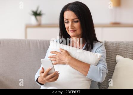 Donna matura sorridente che usa il telefono cellulare, tenendo il cuscino Foto Stock