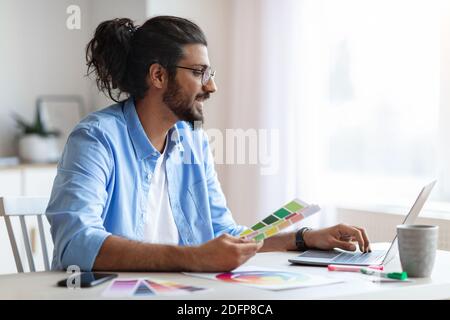 Giovane designer d'interni occidentale che tiene campioni di campioni di campioni di colore e lavora su un computer portatile Foto Stock