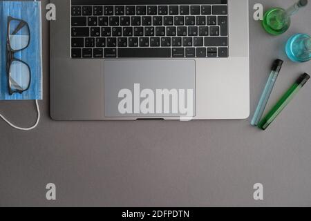 Flatlay di computer portatile, tubi medici e fiasca, maschera e occhiali su una scrivania grigia. Lavorare da casa. Virus e autoisolamento Foto Stock