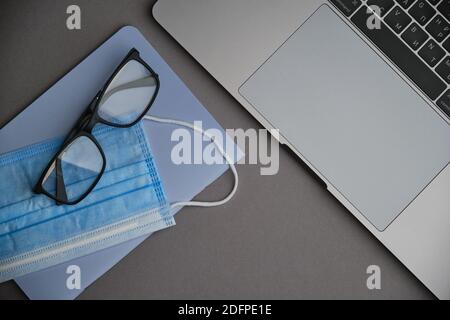 Flatlay di computer portatile, maschera e occhiali su una scrivania grigia. Lavorare da casa. Virus e autoisolamento Foto Stock