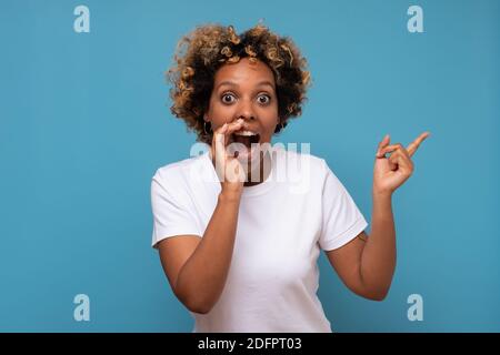 Donna africana con capelli ricci da parte, dicendo segreto. Studio girato su parete blu. Foto Stock