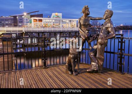 La gente come noi scultura su Mermaid Quay nella baia di Cardiff in una sera di inizio dicembre. La scultura rappresenta una tipica coppia immigrata. Foto Stock