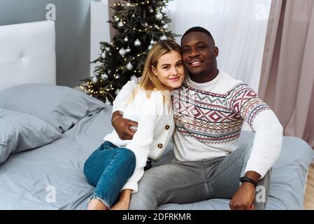 Una coppia multietnica si siede in felpe natalizie sullo sfondo di un albero di Natale, ride e guarda la fotocamera. Marito amorevole e moglie c Foto Stock