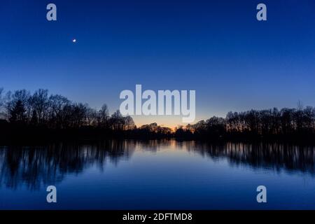 Cielo blu chiaro dopo il tramonto al lago con sagome riflettenti di alberi, venere e luna crescente Foto Stock