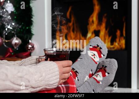 Ragazza che tiene una tazza di tè sulle gambe accanto ad un camino con fuoco e riscalda i piedi. Le gambe della donna sono coperte di coperta e indossare calze decorative di Natale Foto Stock