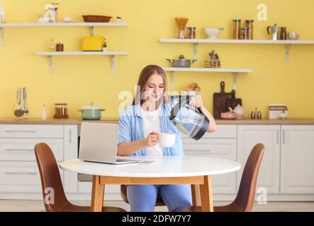 Giovane donna che versa l'acqua bollita dal bollitore elettrico nella tazza in cucina Foto Stock