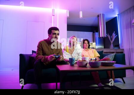 Amico di una giovane coppia viene a casa loro per giocare giochi sulla console di gioco in una piacevole serata. Persone felici che giocano a videogiochi a casa. Festa a casa Foto Stock