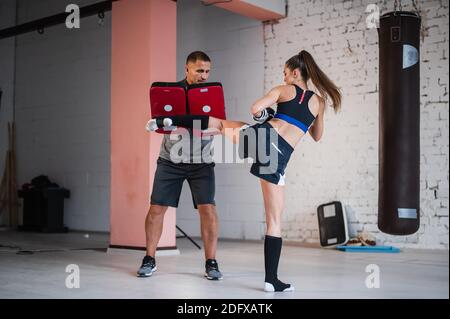 Una giovane ragazza di kickboxer forte calcia il suo partner scintillante con una zampa di pugilato in una sessione di allenamento personale Foto Stock