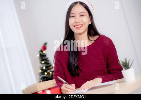 Giovane donna asiatica che scrive cartolina nel giorno di Natale a casa, vigilia e celebrare, femmina che scrive messaggio sul biglietto di auguri con dare scatola regalo, vacanza Foto Stock