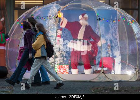 Seattle, Washington, Stati Uniti. 6 dicembre 2020. DaN Kemmis, noto come Seattle Santa, si estende come un gruppo di giovani donne che cammina accanto alla sua bolla protettiva in plastica nel quartiere Greenwood di Seattle. Incapace di programmare eventi privati a causa della pandemia del coronavirus, Kemmis sta facendo apparizioni pubbliche fisicamente distanziate con alcune donazioni che avvantaggiano i rifugi locali dei senzatetto. Credit: Paul Christian Gordon/Alamy Live News Foto Stock