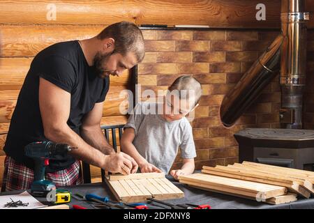 Papà e figlio stanno lavorando su un prodotto in legno, realizzando contrassegni per il fissaggio, gli attrezzi e il legno sul tavolo in officina. Concetto di formazione di carpenteria Foto Stock