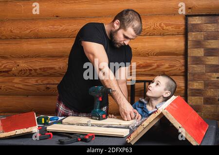 Papà e figlio stanno lavorando su un prodotto in legno, facendo marcature per il fissaggio, utensili e legno sul tavolo. Concetto di formazione di carpenteria per bambini Foto Stock