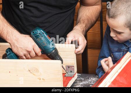 Padre e figlio insieme fanno una casa di uccelli in legno nel laboratorio. Padre allegro con un ragazzino che fora una tavola di legno usando un cacciavite su un woo Foto Stock