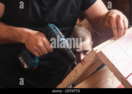 Padre e figlio insieme fanno una casa di uccelli in legno nel laboratorio. Padre allegro con un ragazzino che fora una tavola di legno usando un cacciavite su un woo Foto Stock