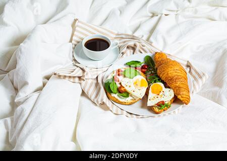 Concetto di San Valentino. Colazione a letto il giorno di San Valentino. Panini con croissant, bagel, formaggio spalmabile e uova fritte su un letto bianco bac Foto Stock