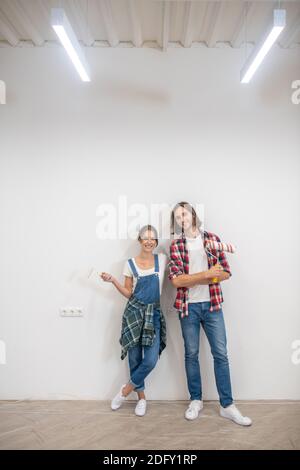 Giovane coppia in piedi vicino al muro con rulli di vernice e. mani Foto Stock