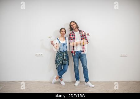 Giovane coppia in piedi vicino al muro con rulli di vernice e. mani Foto Stock