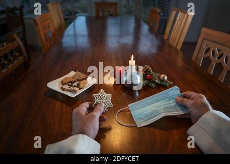 Mani di un uomo anziano che tiene un biscotto di Natale e una maschera chirurgica contro l'infezione da covid-19 ad un grande tavolo di famiglia con sedie vuote, solo Foto Stock