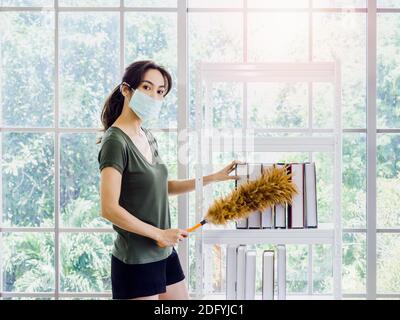 Giovane bella donna asiatica, casalinga in panno casual che indossa maschera protettiva pulizia libri su libreria con piumino in soggiorno su Foto Stock