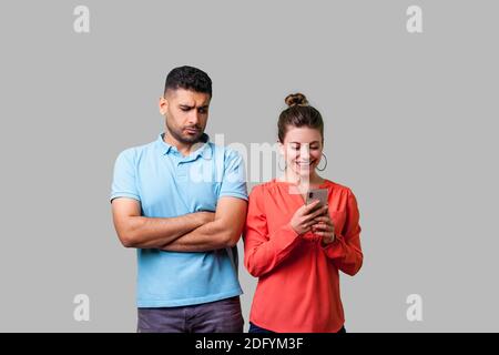 Ritratto di geloso fidanzato in piedi con le braccia conserte, cercando di sospetto a donna barare usando il telefono e sorridente felicemente, relazioni infedele. Foto Stock