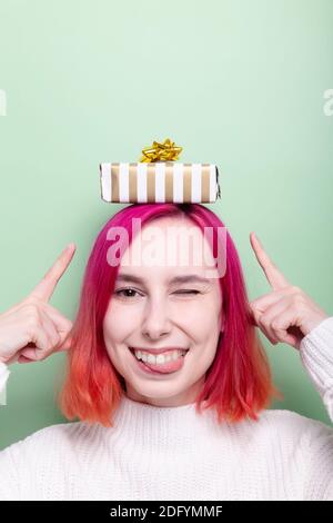 Donna in pullover bianco con confezione regalo sulla testa fa il viso divertente e punta le dita in regalo Foto Stock