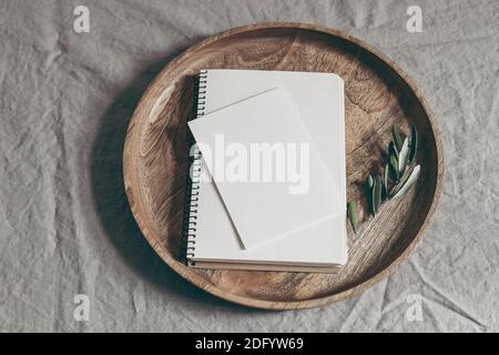 Moody femminile fedding stationery mock-up scena. Biglietto d'auguri vuoto, blocco note e ramo verde oliva su vassoio di legno. In alto Foto Stock