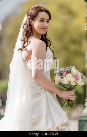 Sposa in abito da sposa guardando e sorridendo alla macchina fotografica primo piano di sposa con bouquet di sposa all'esterno con sfocato sfondo Foto Stock