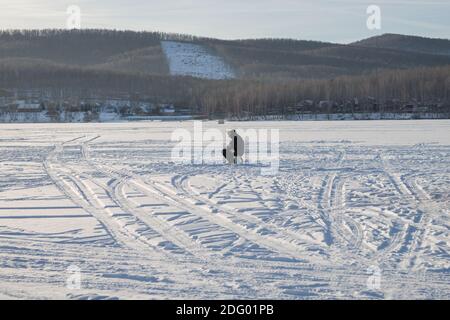 Il pescatore si siede e cattura il pesce sul lago innevato, in serata Foto Stock