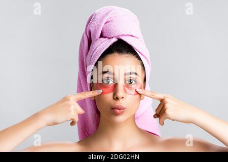 bella donna con asciugamano sui capelli che punta all'occhio di idrogel macchie sulla faccia isolate su grigio Foto Stock