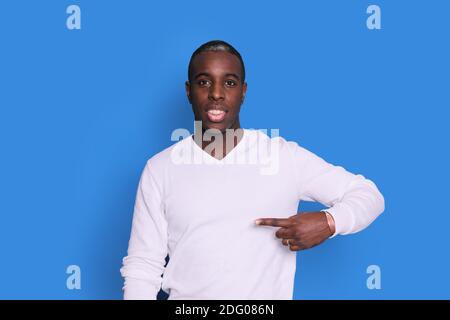 Foto interna di un giovane afro-americano raffigurato isolato su sfondo blu che punta al suo maglione bianco che attira l'attenzione alla pubblicità su Foto Stock