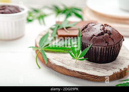 Muffin alle erbe infestate con cannabis sulla parte superiore, foglie di cannabis, rami di canapa sulla tavola bianca. Muffin con cupcake di cioccolato alla marijuana e cbd con erbacce. Marijuana medica Foto Stock