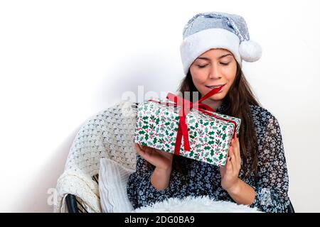 giovane brunetta in un cappello babbo natale in argento seduto su una sedia e tenendo una scatola regalo in mano, isolato su uno sfondo bianco Foto Stock