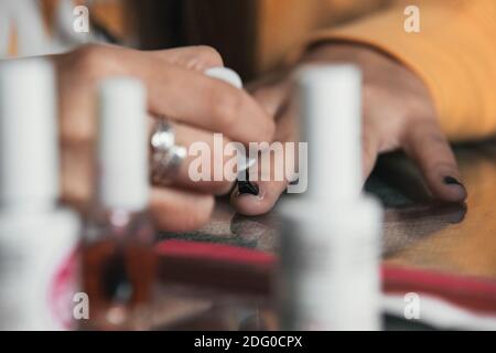 pittura chiodi blackclose-up colpo di una mano bianca in una donna una giacca arancione dipinge i suoi chiodi neri su un bicchiere tavolo con lattine di vernice nello straniero Foto Stock